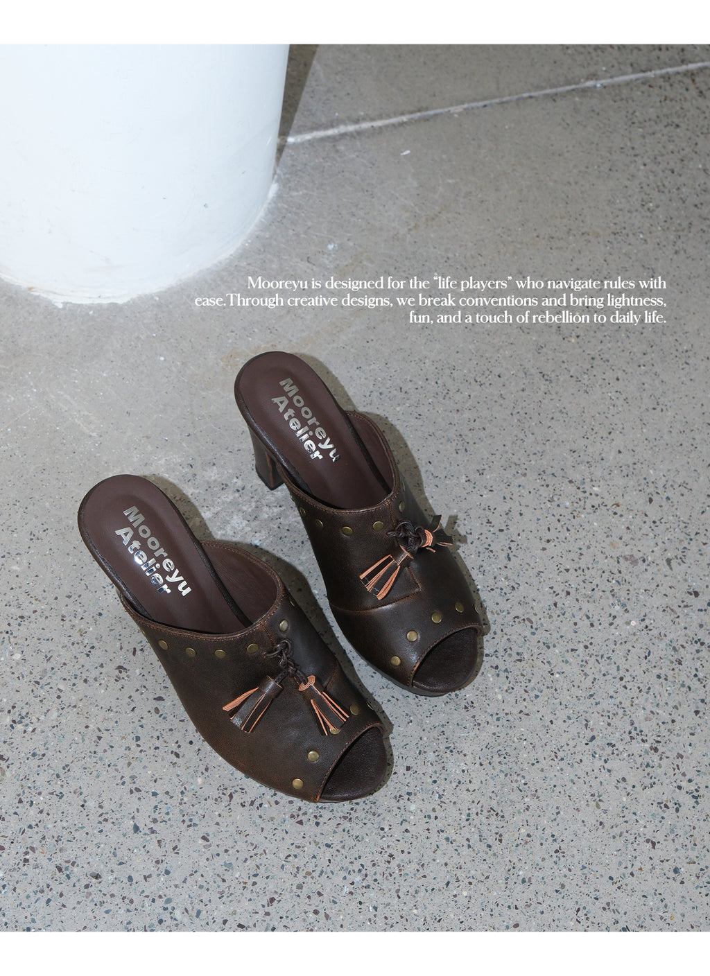 Top-down overhead shot of two brown nomad leather mule clogs with studs and tassels placed on speckled concrete floor