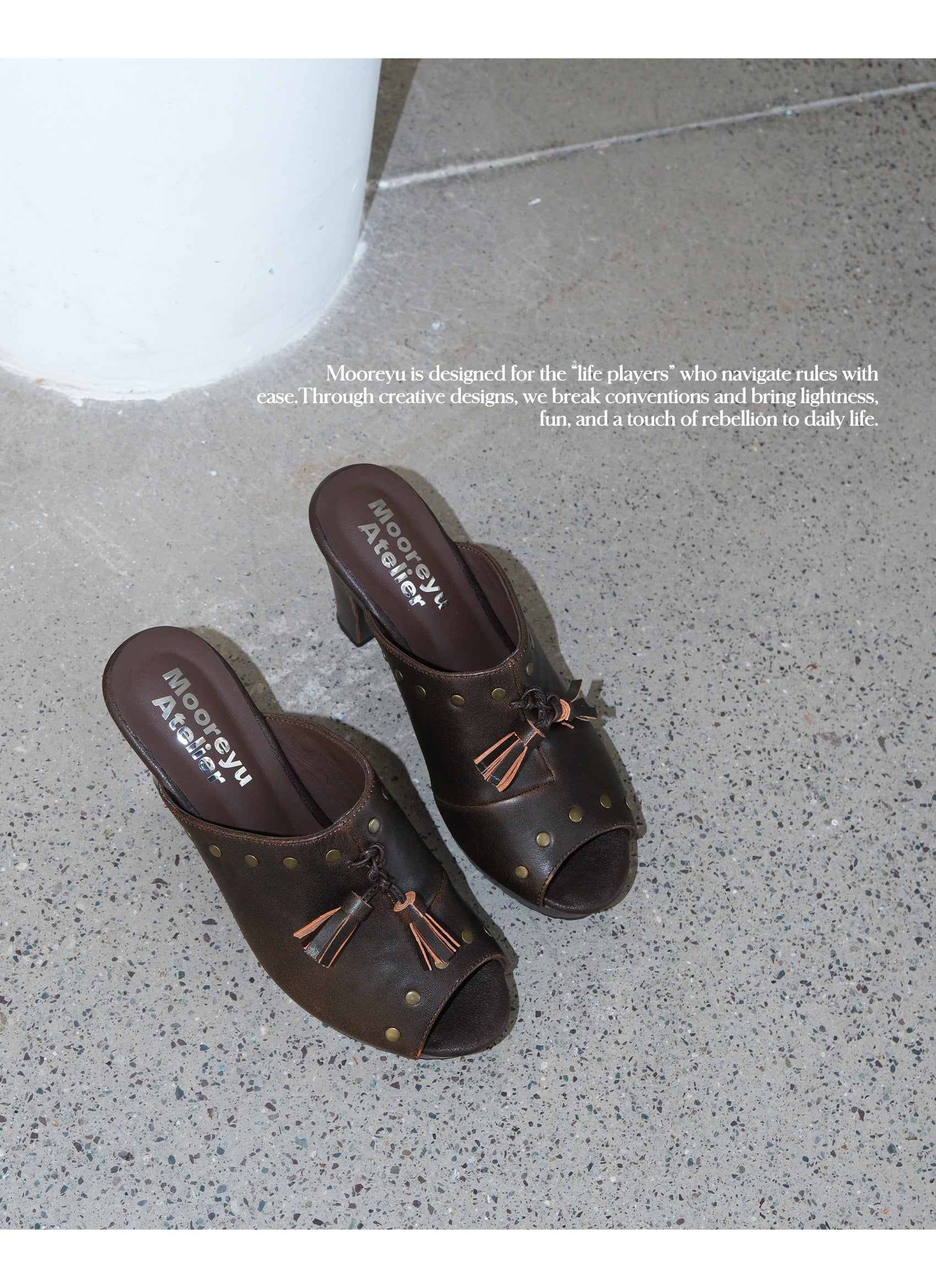 Top-down overhead shot of two brown nomad leather mule clogs with studs and tassels placed on speckled concrete floor