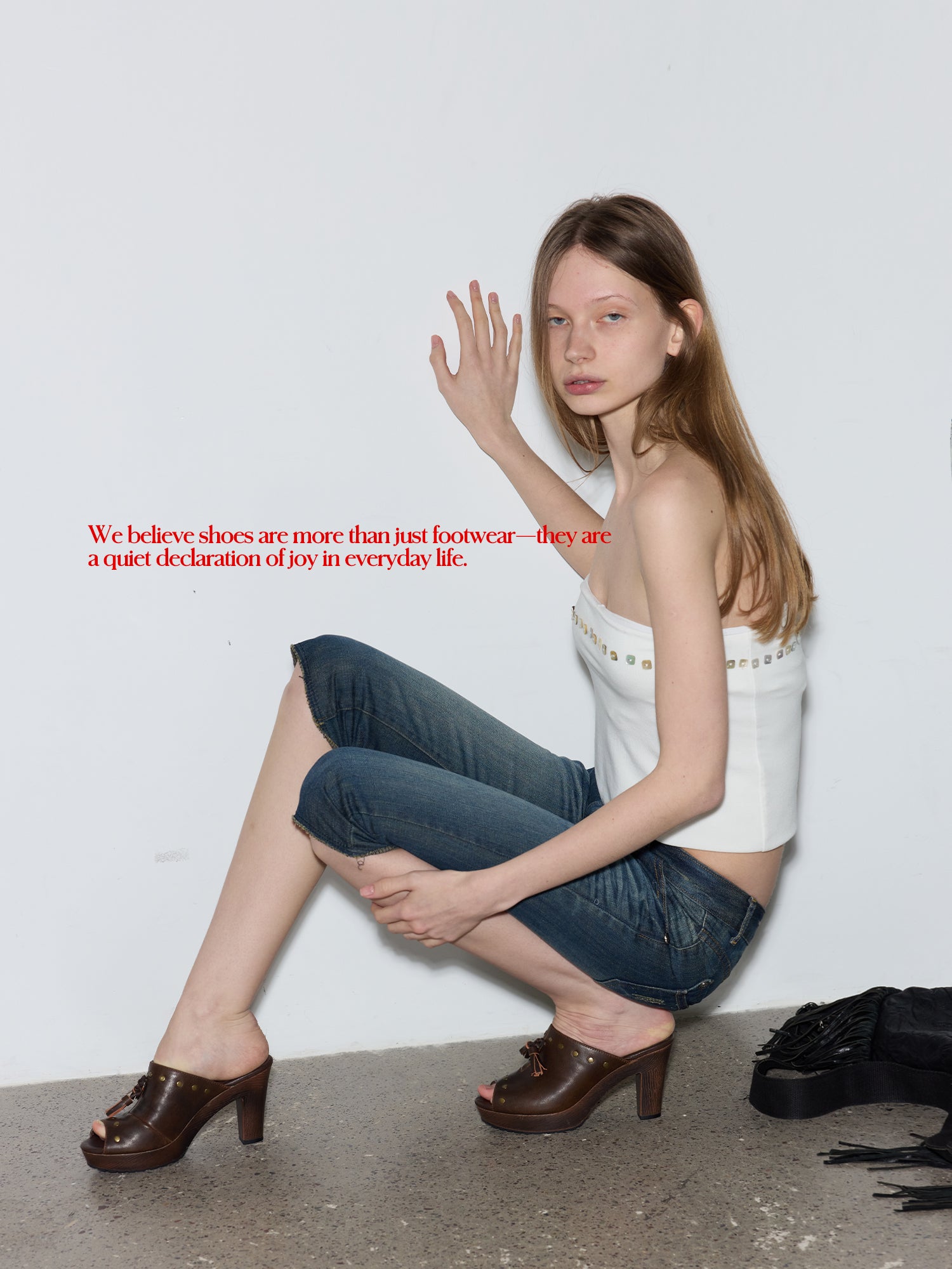 Female model squatting against white wall wearing white strapless top & denim capris, styled with distressed brown studded tassel wood block mule slides