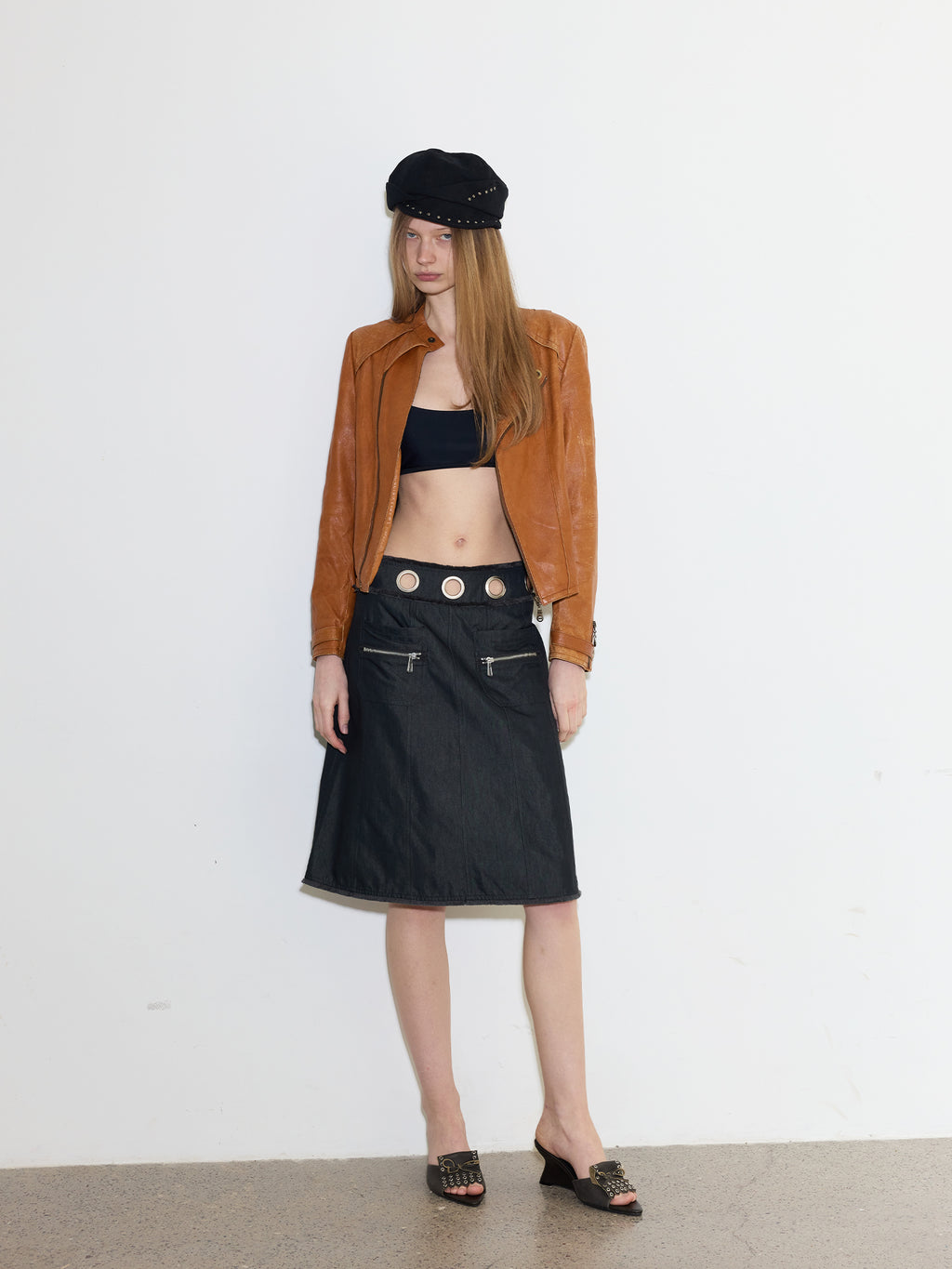 Full body shot of female model in vintage brown leather jacket, black crop top & denim midi skirt with metal eyelet waistband, wearing dark grey eyelet bow leather wedge mule slides