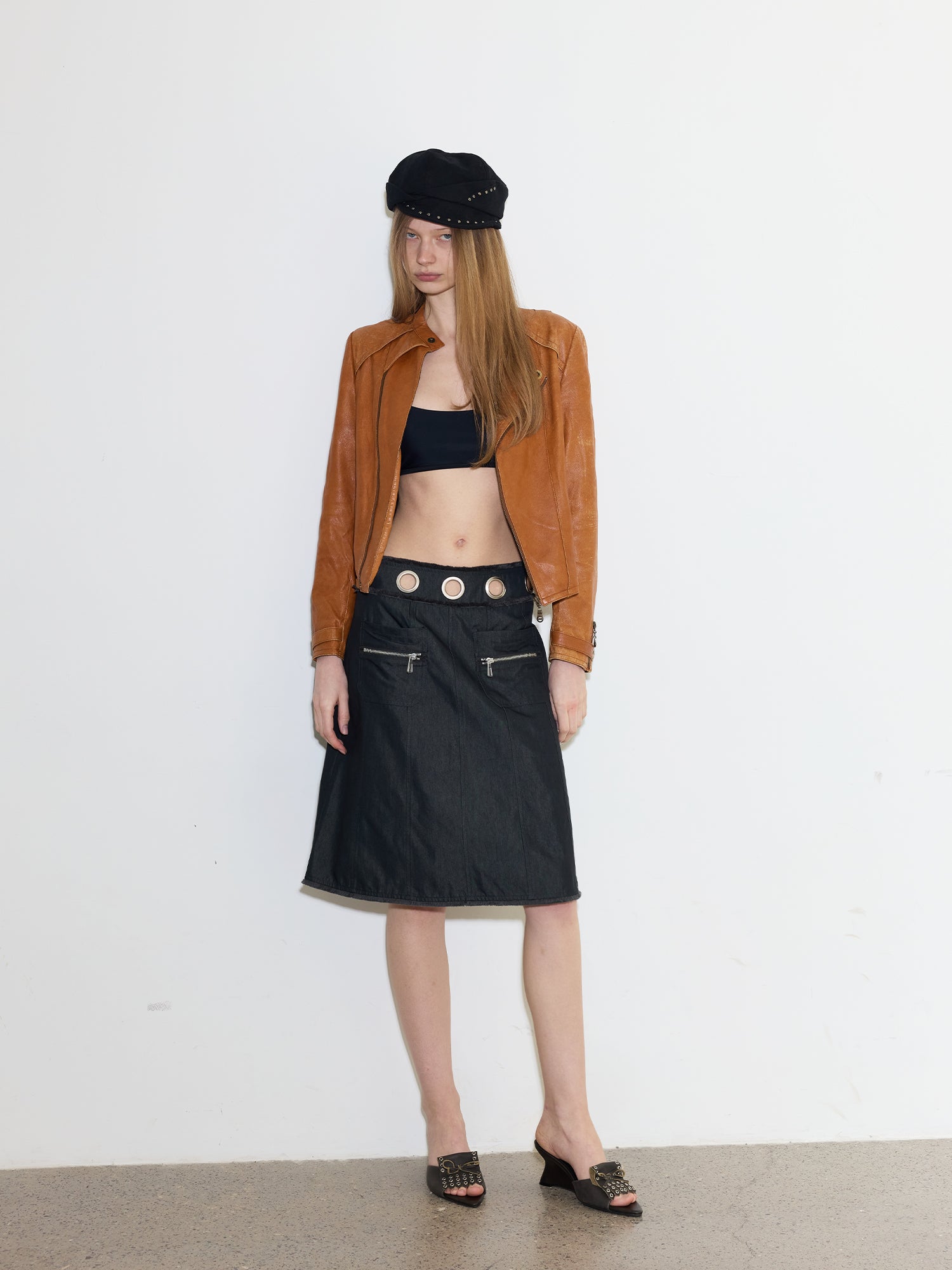 Full body shot of female model in vintage brown leather jacket, black crop top & denim midi skirt with metal eyelet waistband, wearing dark grey eyelet bow leather wedge mule slides