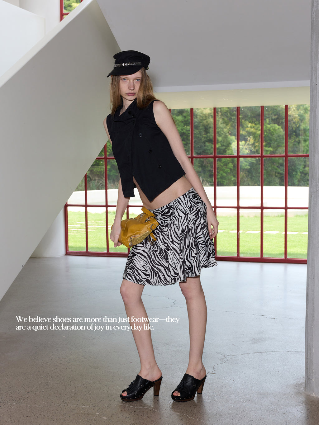 Full body female model wearing black sleeveless vest, zebra print mini skirt, black stud cap & yellow leather handbag, styled with black pebbled leather fringe wood platform mule clogs