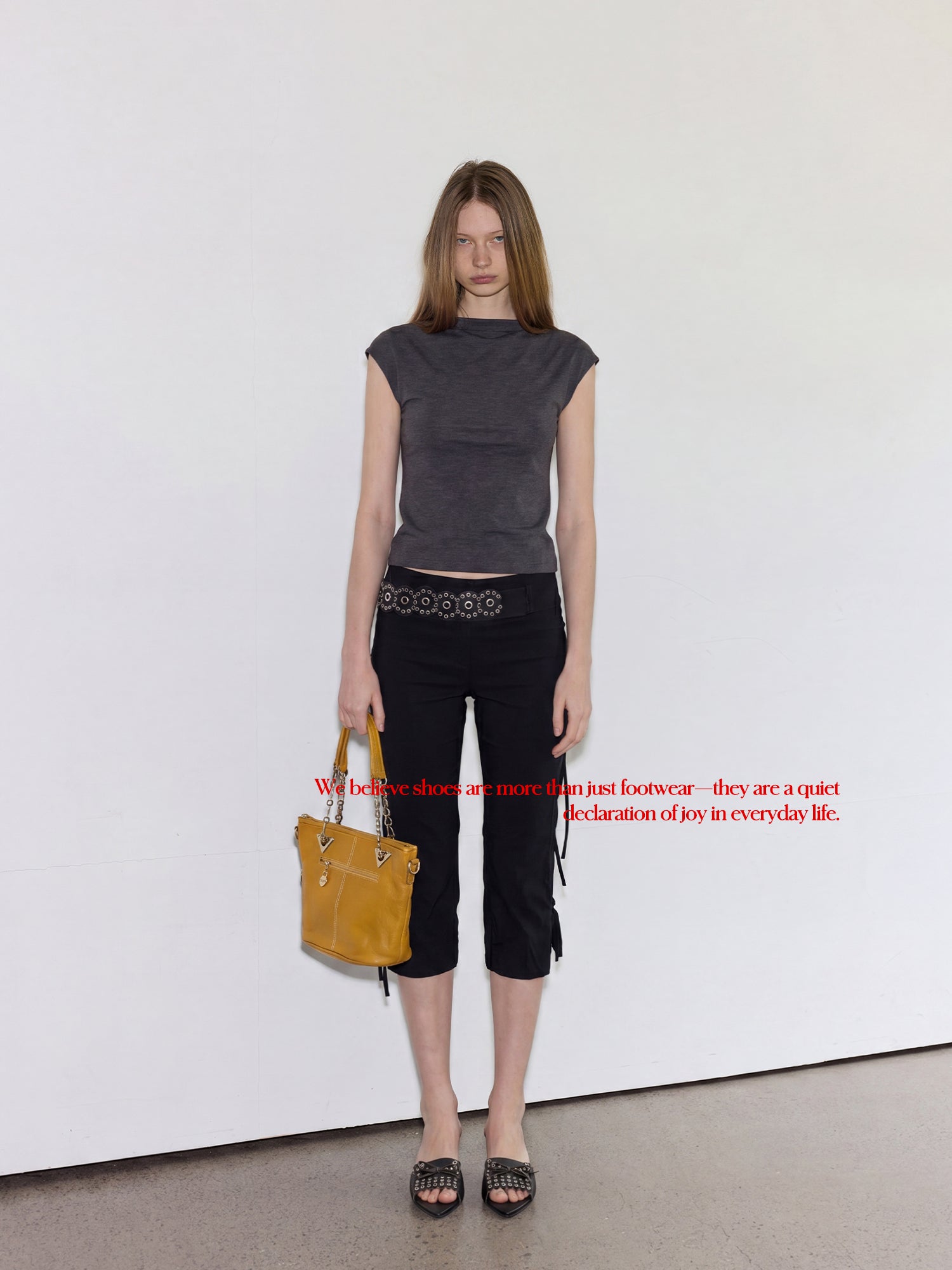 Full length studio shot of female model in grey cap-sleeve top & black cropped capri pants, holding yellow leather bucket bag, wearing vintage dark grey eyelet bow wedge leather mule slides