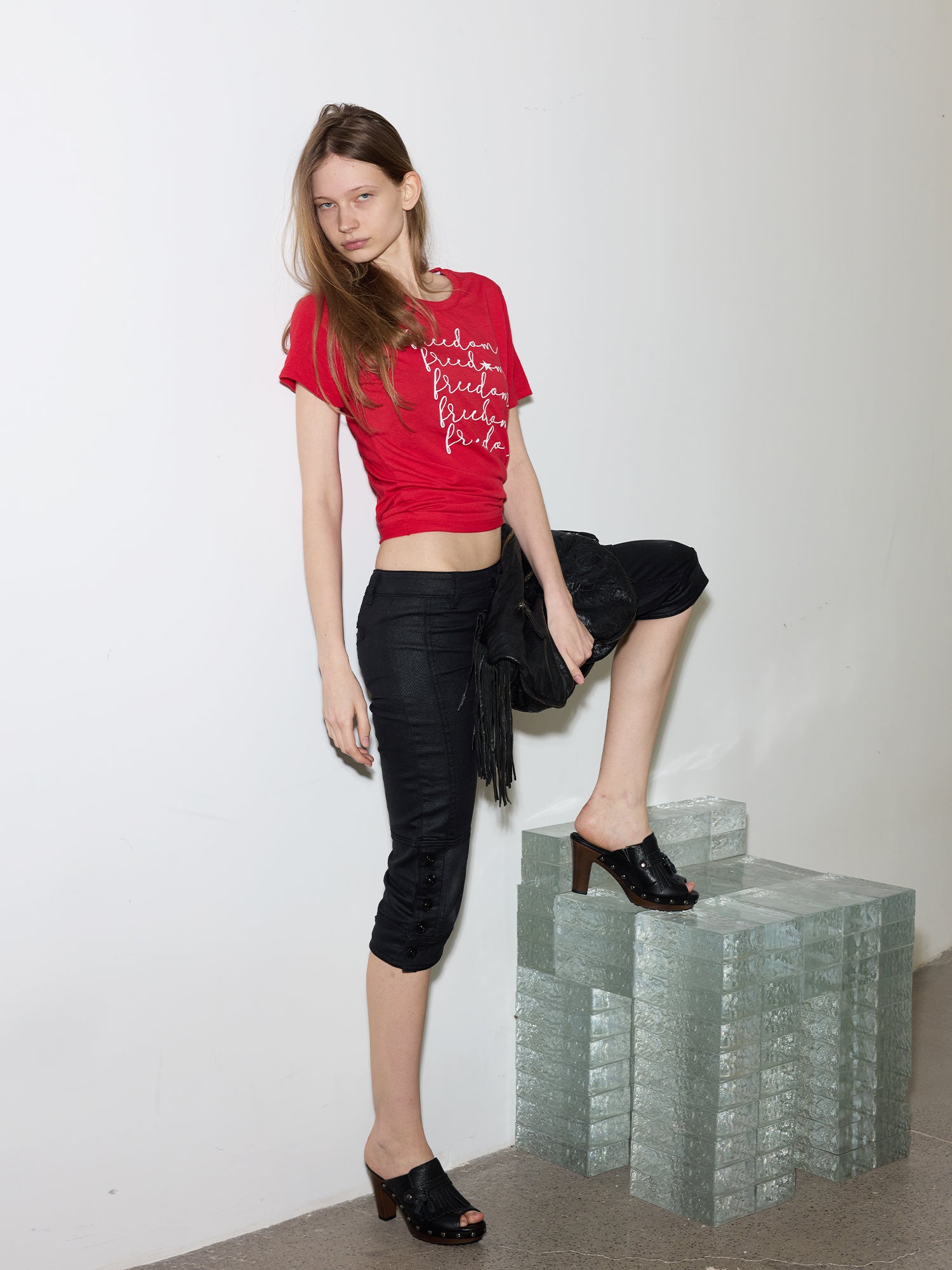 Model leaning against plain white wall in cropped red graphic tee & black capri jeans, one foot on glass block steps, wearing black fringe stud leather wood grain mule clogs