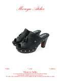 White background MOOREYU ATELIER solid black pebbled full grain leather mule clogs, peep toe leather fringe tassels & surrounding silver studs, 10cm faux wood grain platform block heels studio shot
