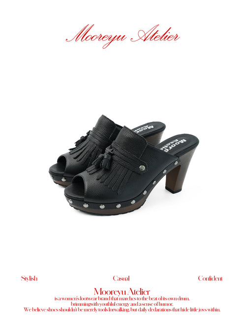 White background MOOREYU ATELIER solid black pebbled full grain leather mule clogs, peep toe leather fringe tassels & surrounding silver studs, 10cm faux wood grain platform block heels studio shot