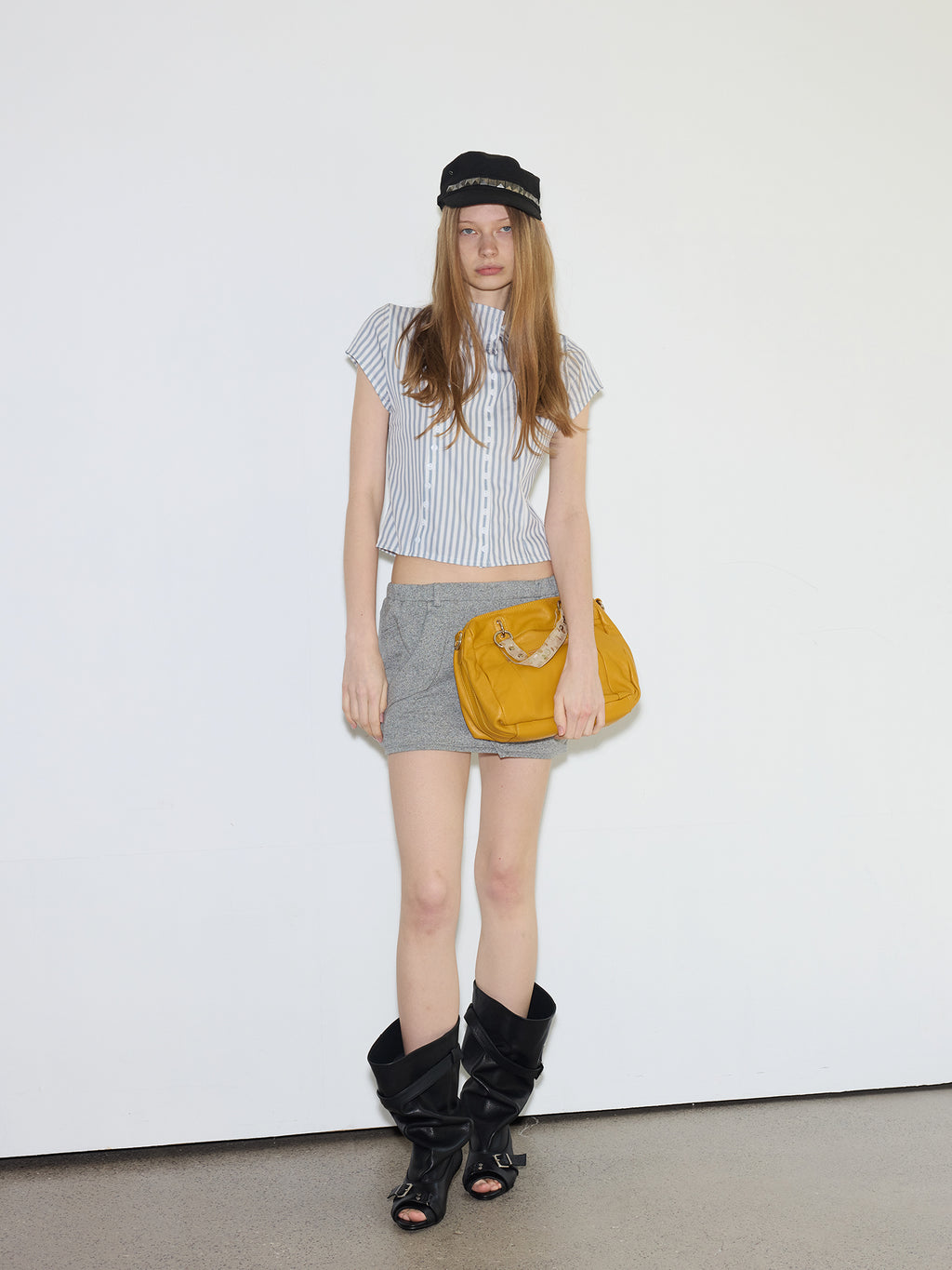 Full-body shot of female model wearing striped cropped shirt and grey mini skirt, styled with MOOREYU ATELIER black peep-toe slouch mid-calf leather boots with dual buckle straps, studio white wall background