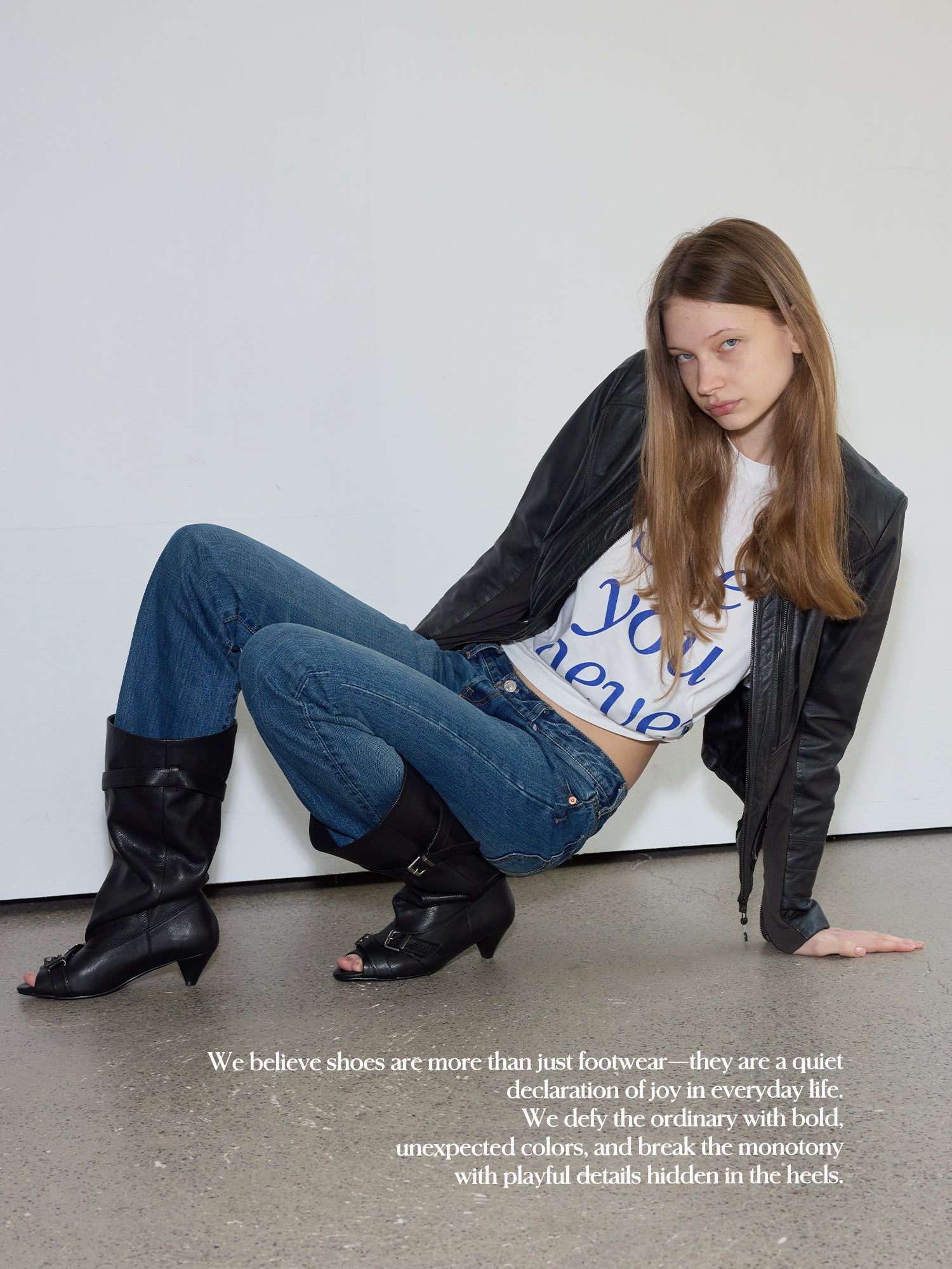 Model crouching outfit photo, black leather jacket, vintage blue jeans, wearing black peep toe slouch mid calf leather buckle boots, minimalist concrete studio with brand quote text overlay