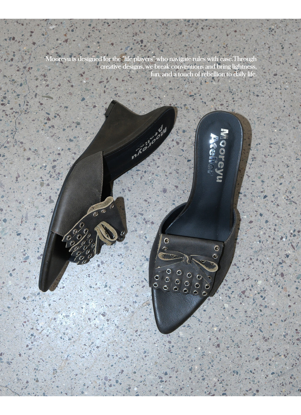 Top-down overhead shot of two dark grey wasteland leather wedge mule slides on speckled concrete floor, highlighting metal eyelet fringe bow decor and branded sheepskin insole print