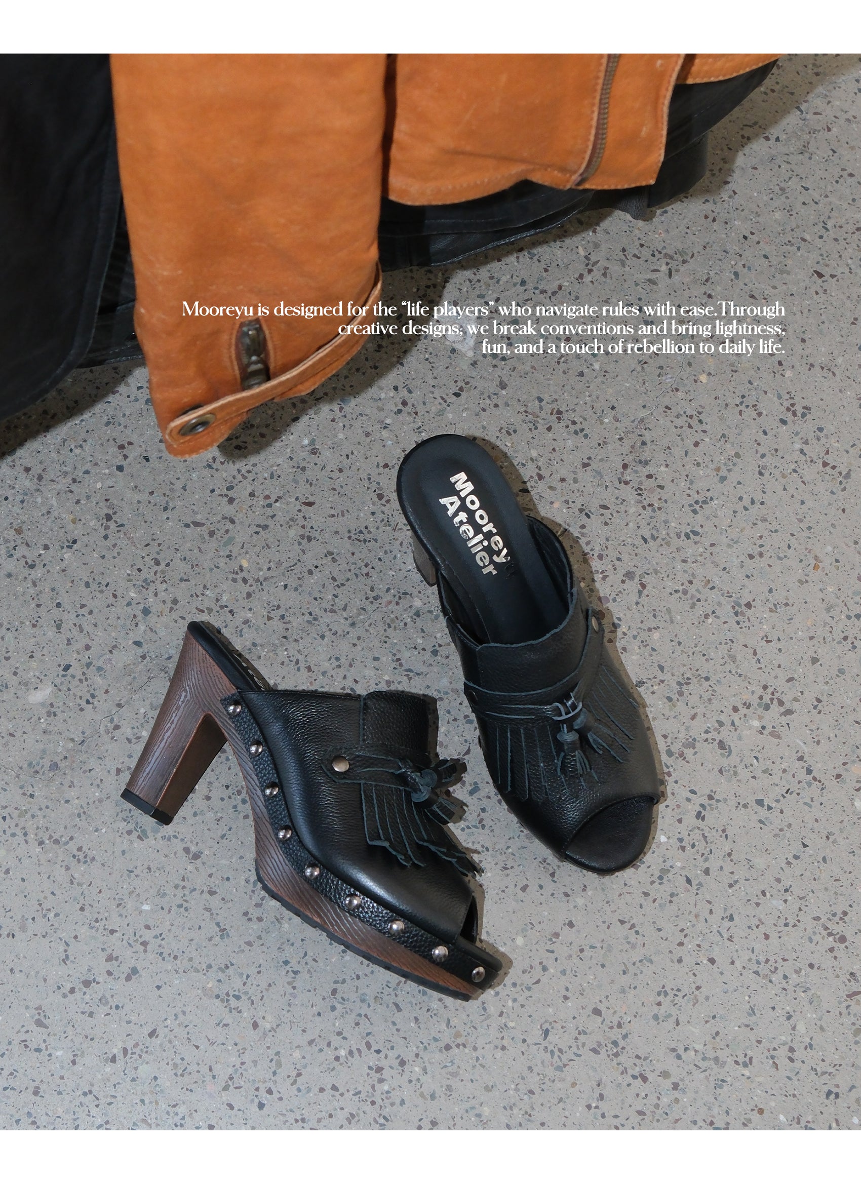 Two black boho fringe leather mule clogs laid sideways on speckled concrete floor, branded sheepskin insole print, wood grain heel and surrounding silver metal studs detail