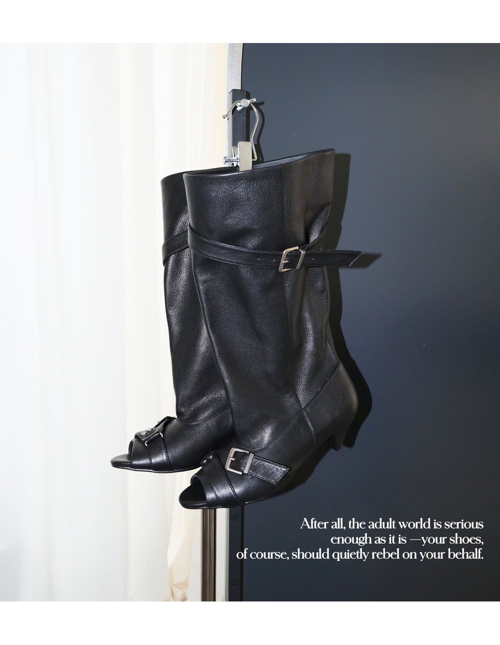 Hanging product shot of MOOREYU ATELIER black full grain leather peep toe slouch mid calf buckle boots, display hanger, dark grey backdrop with brand text quote overlay