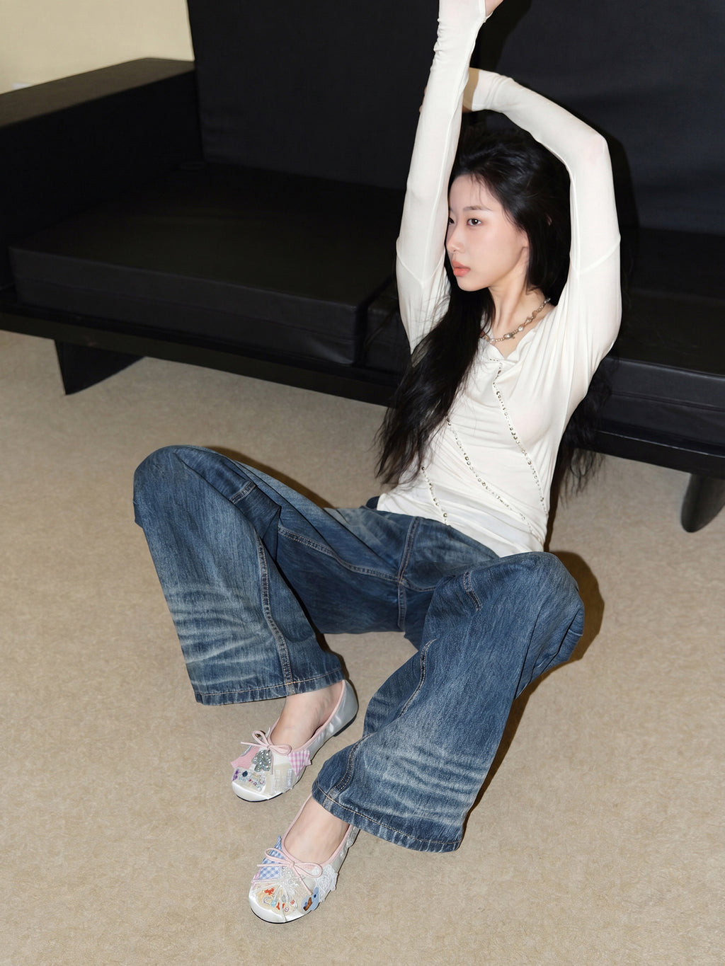 Female model sitting on carpet with arms raised, wearing vintage heart patch satin ballet flats, casual coquette streetwear look